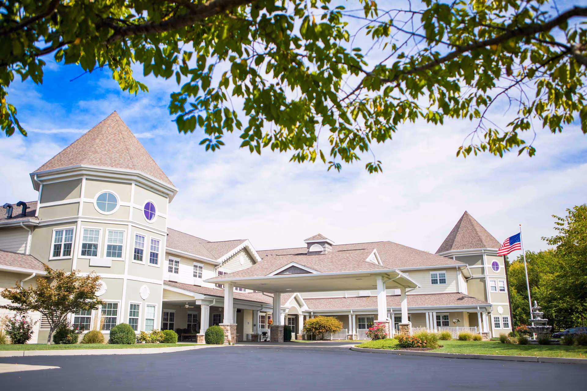 Exterior view of a senior living facility with two turret-style towers, a covered entrance, well-maintained landscaping, and an American flag on a flagpole. The sky is partly cloudy and tree branches frame the top of the image.