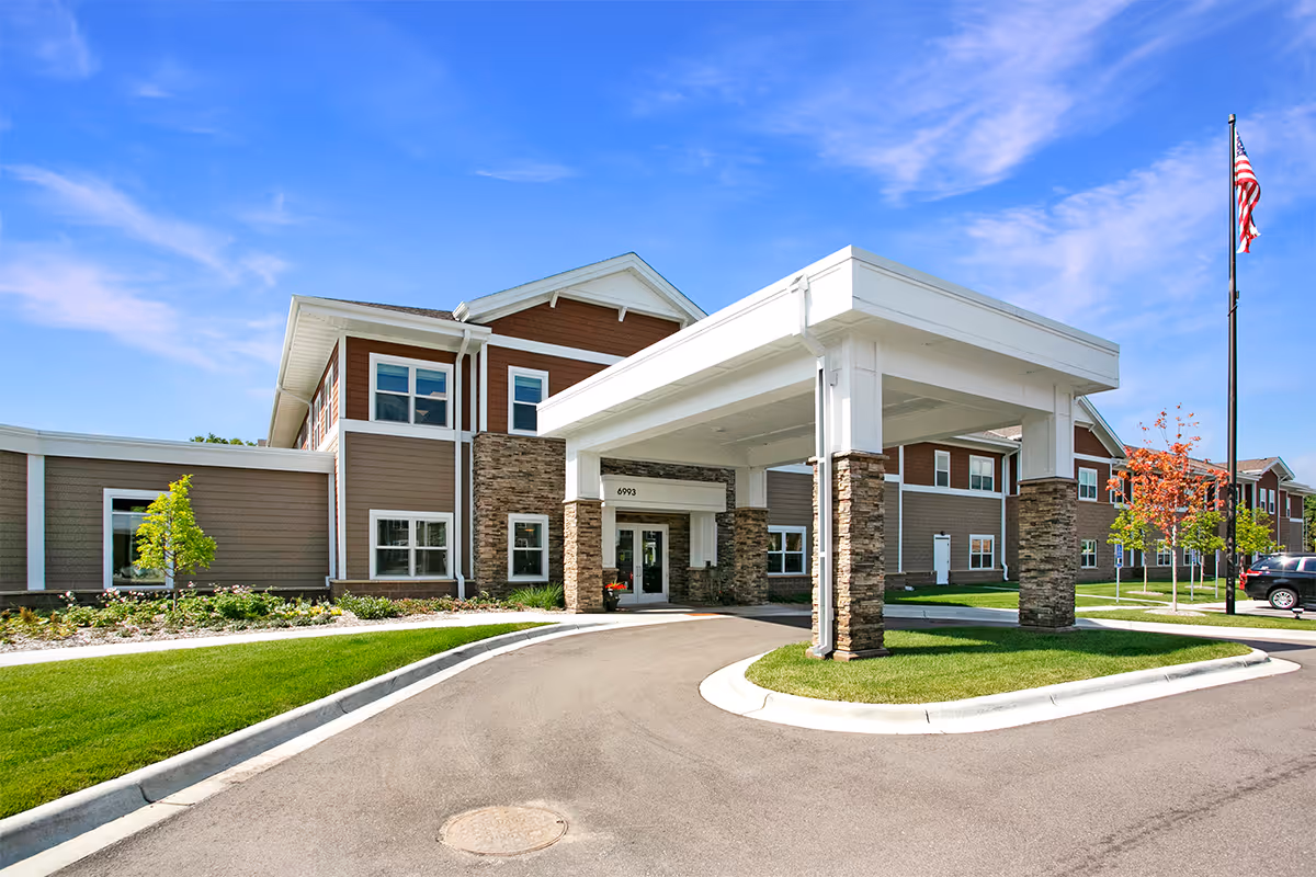Front entrance of a two-story senior living facility with a covered porte-cochere, driveway and landscaped lawn under a blue sky.