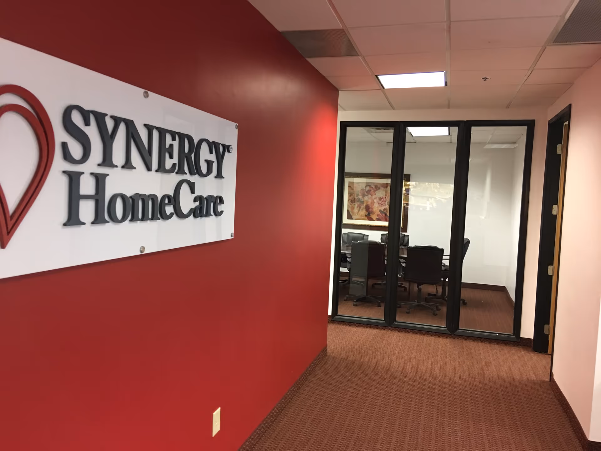 Hallway with Synergy HomeCare sign on red wall and glass-walled conference room.