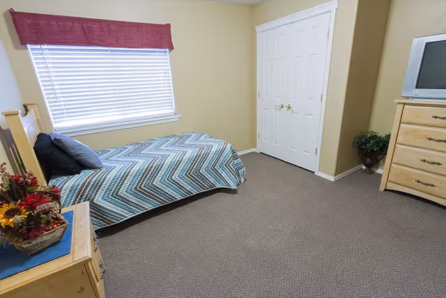 A simple bedroom with a single bed covered in a blue and gray chevron patterned blanket, two pillows, a wooden nightstand with a floral arrangement, a window with closed blinds and a red valance, a double-door closet, a wooden dresser with a small TV on top, and a potted plant in the corner.