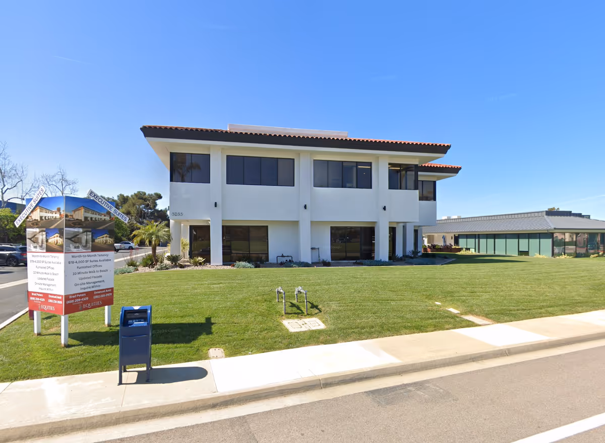 Two-story white building with manicured lawn, signage board, and a sidewalk in front.