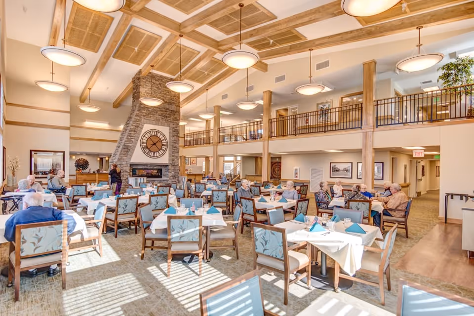 Bright two-story dining room with many set tables, a stone fireplace topped by a large clock, and residents seated at tables.