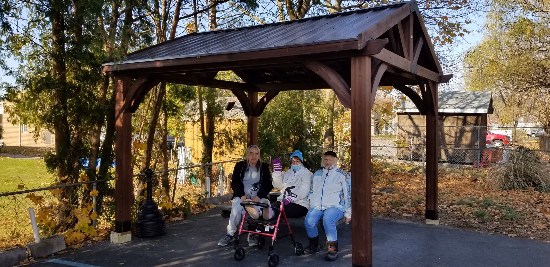 Three people sit on a bench beneath a wooden gazebo in an outdoor senior living courtyard.