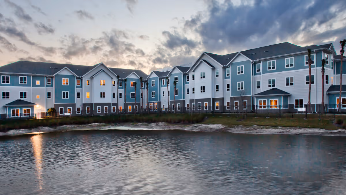 Three-story residential building with lit windows facing a pond at dusk.