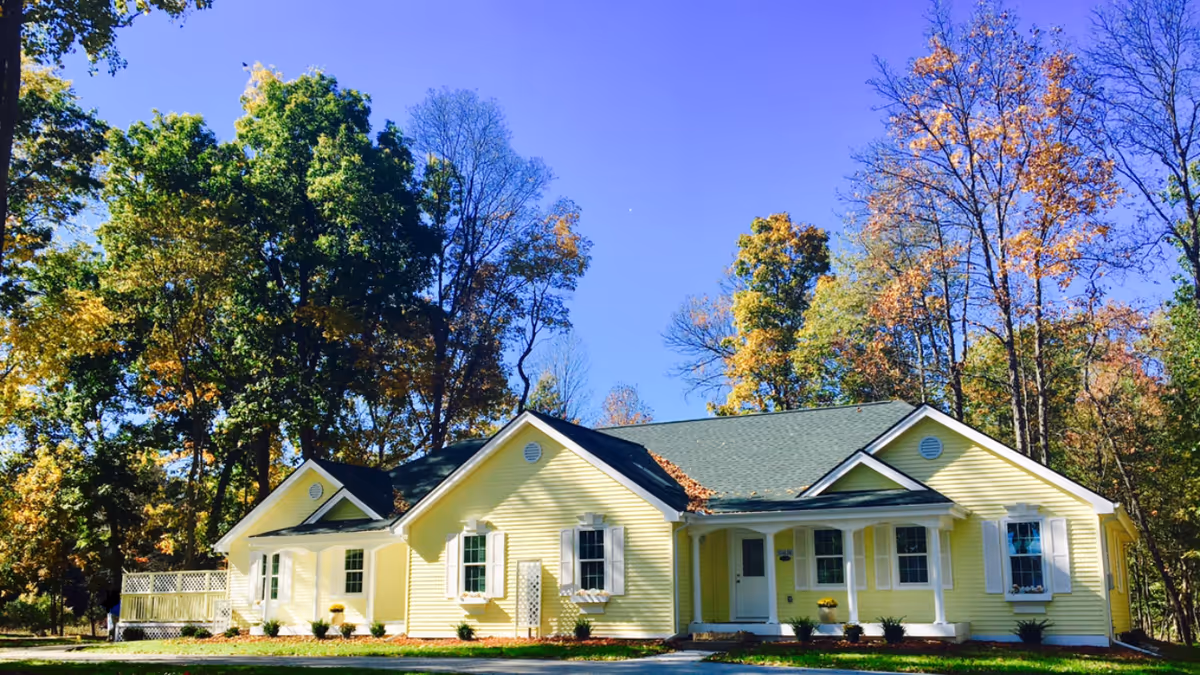 Front view of a yellow single-story senior living building surrounded by trees under a clear blue sky.