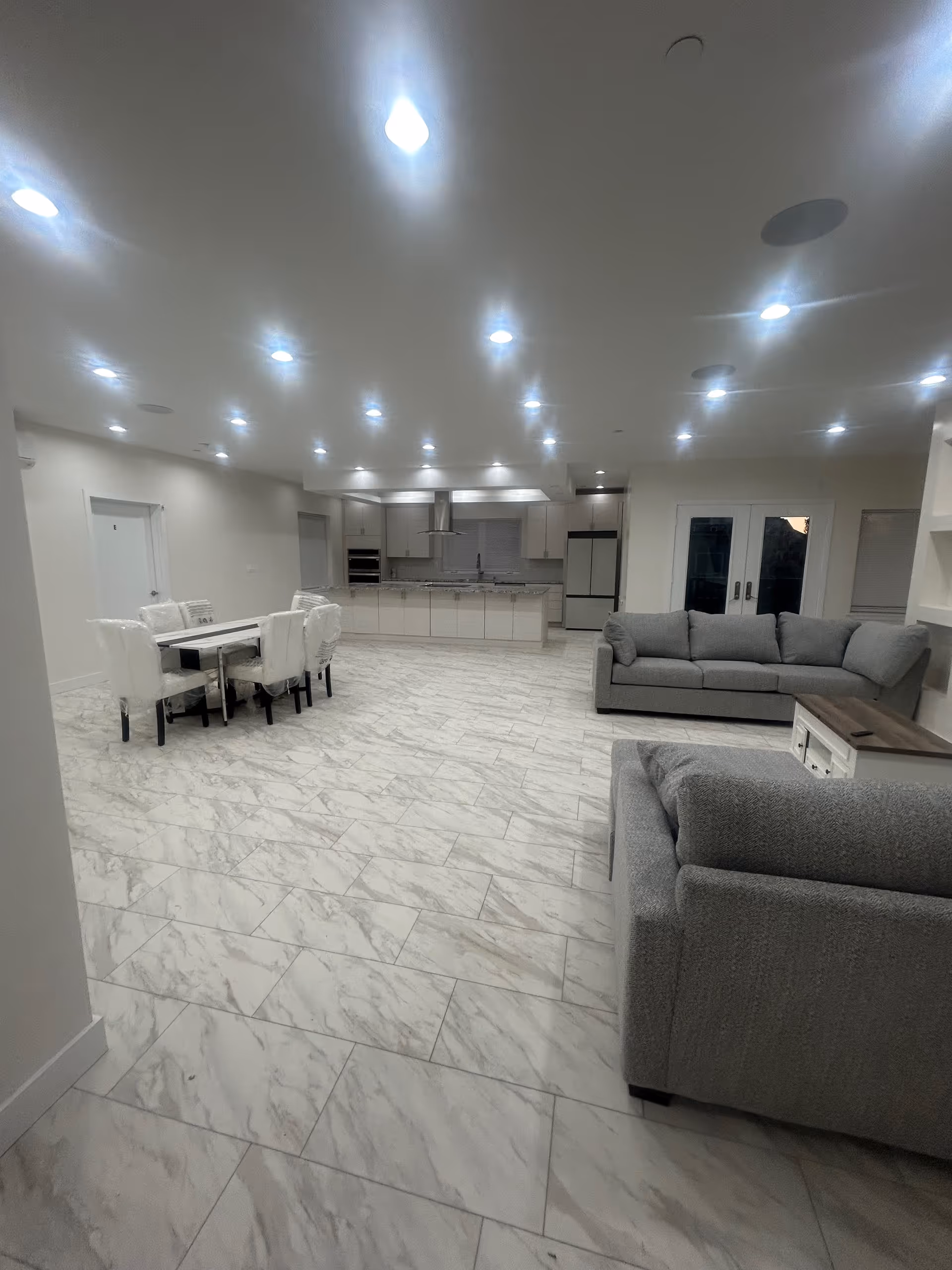 Bright open common area with white marble floors, kitchen, dining table, and gray sofas.