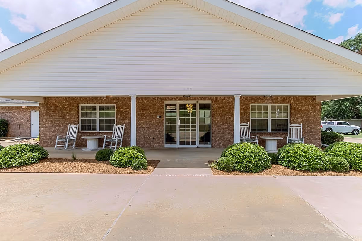 Front exterior view of a senior living facility with a covered entrance supported by white columns. There are two sets of white rocking chairs and small round tables on either side of the glass double doors. The building has brick walls and white siding on the upper part. Green bushes and mulch line the front of the building, and a concrete driveway is visible in the foreground under a partly cloudy sky.