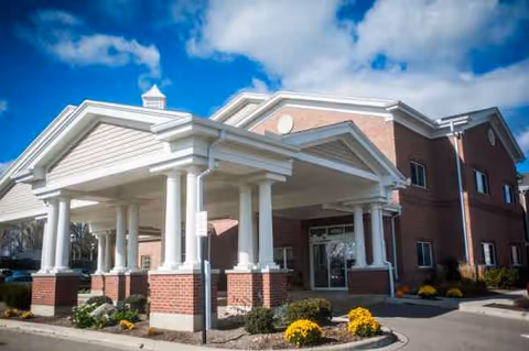 Brick senior living facility entrance with white columns and a covered porch under a blue sky.