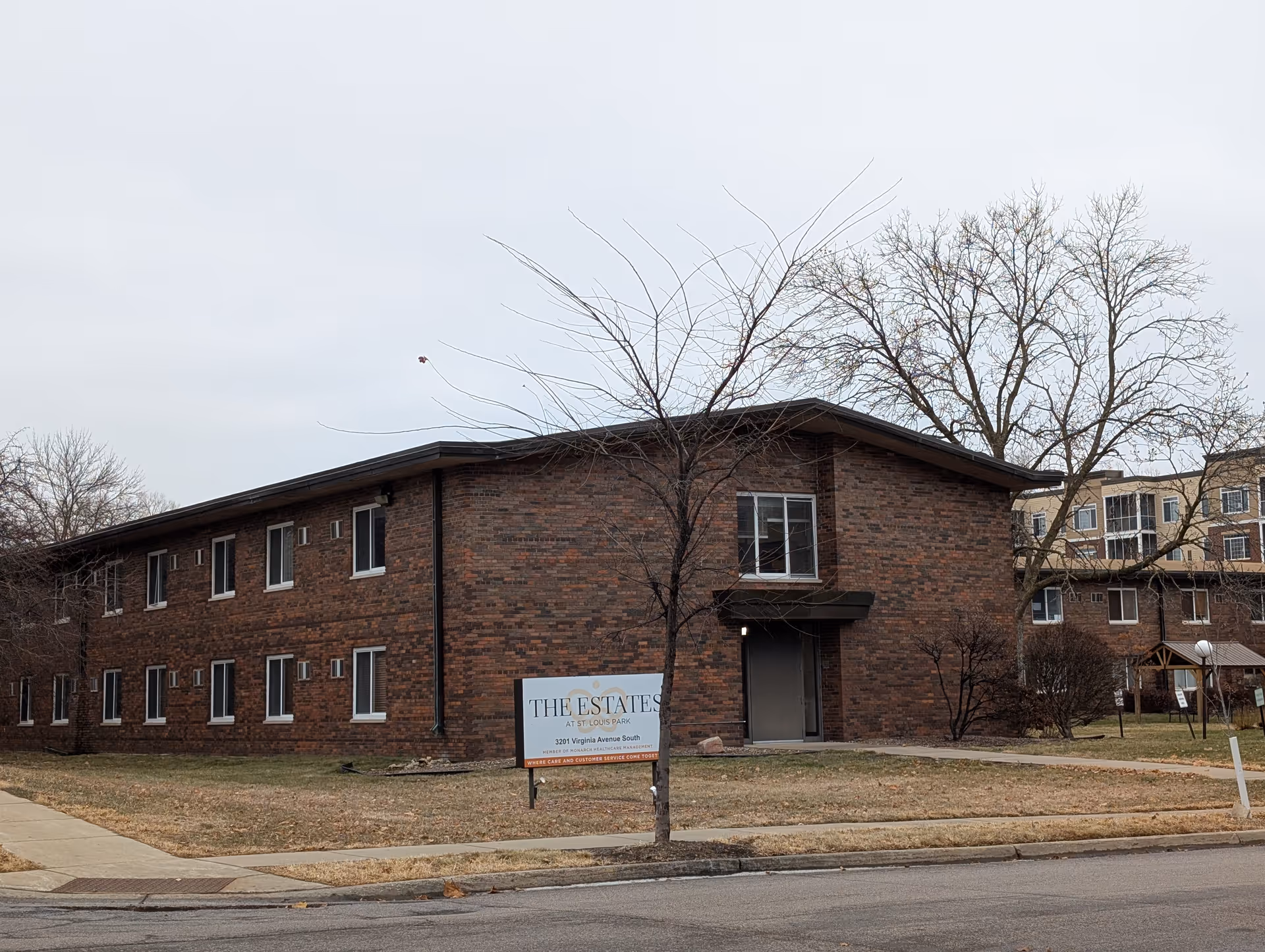 Two-story brick building with leafless trees and a lawn, a sign in front reading "The Estates" on a corner lot.