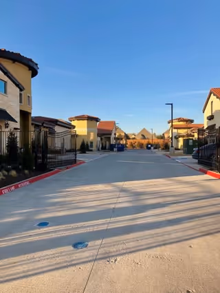 Gated driveway entrance lined by Mediterranean-style senior living buildings under a clear blue sky.