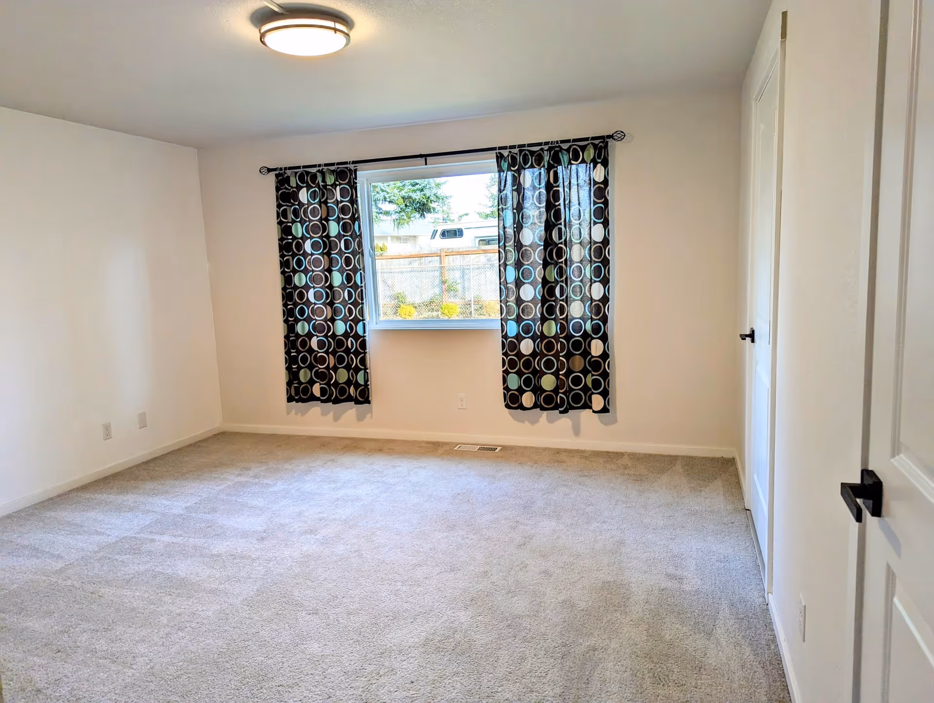 Empty bedroom with light gray carpet, white walls, a window with patterned curtains, and a ceiling light fixture. There is a door with a black handle on the right side of the room.