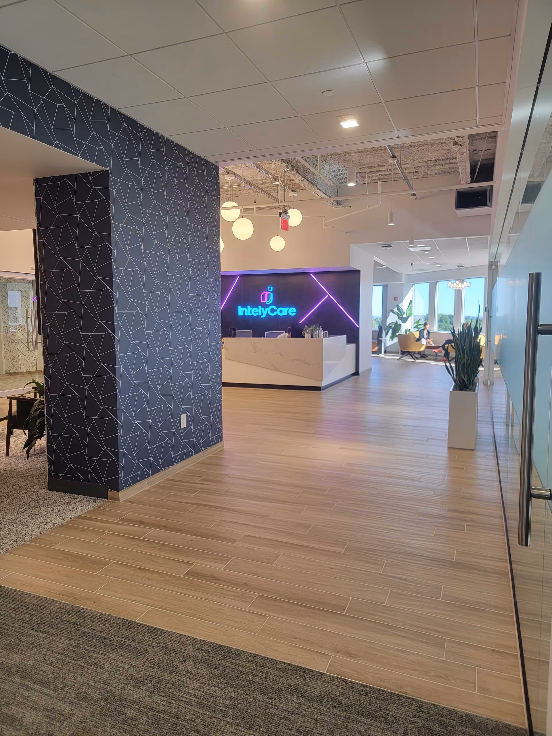A modern lobby features a reception desk, neon IntelyCare sign, blue geometric wall, and seating.