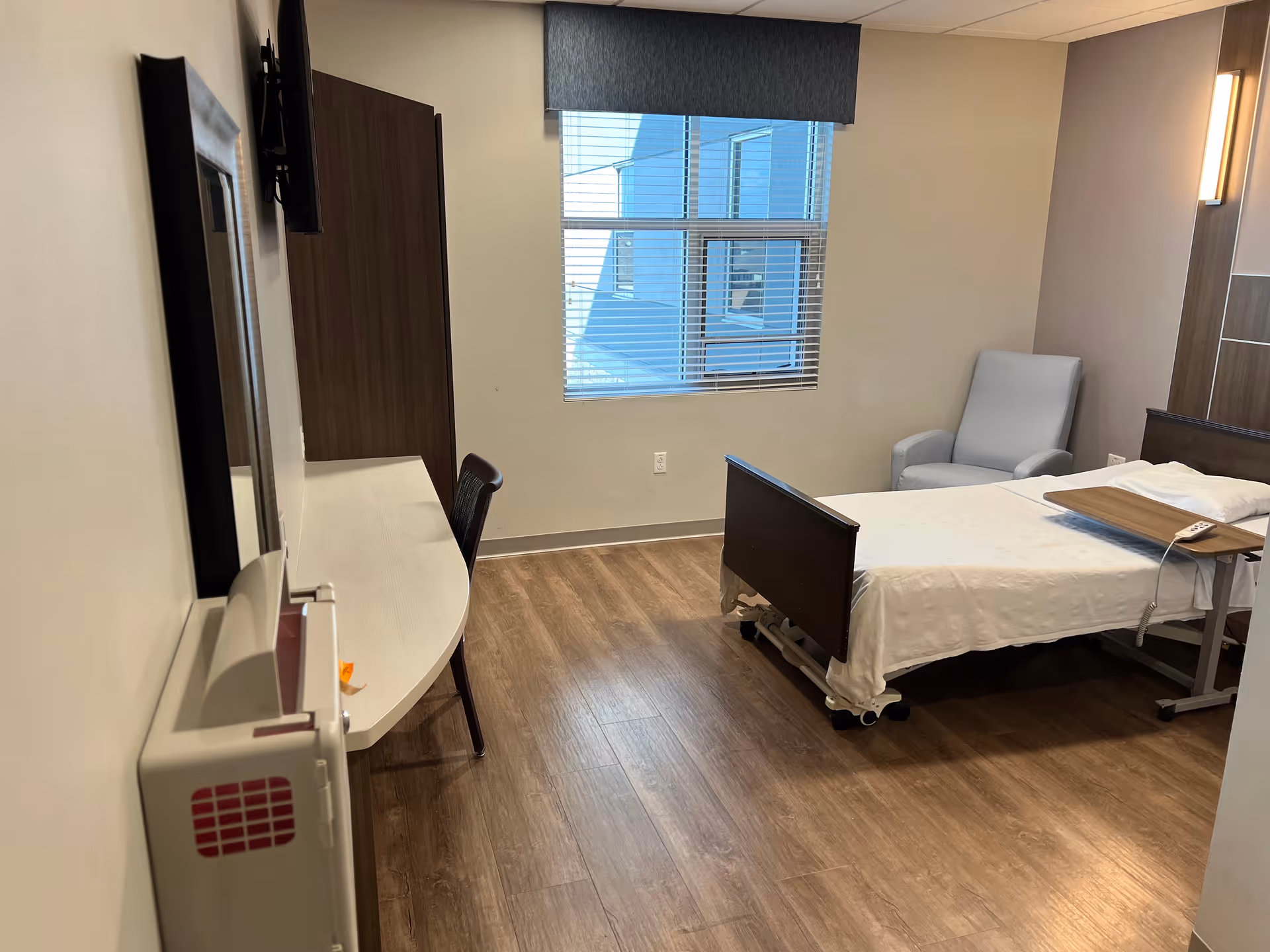 Private bedroom in a senior living facility with a bed, chair, and desk by a window.
