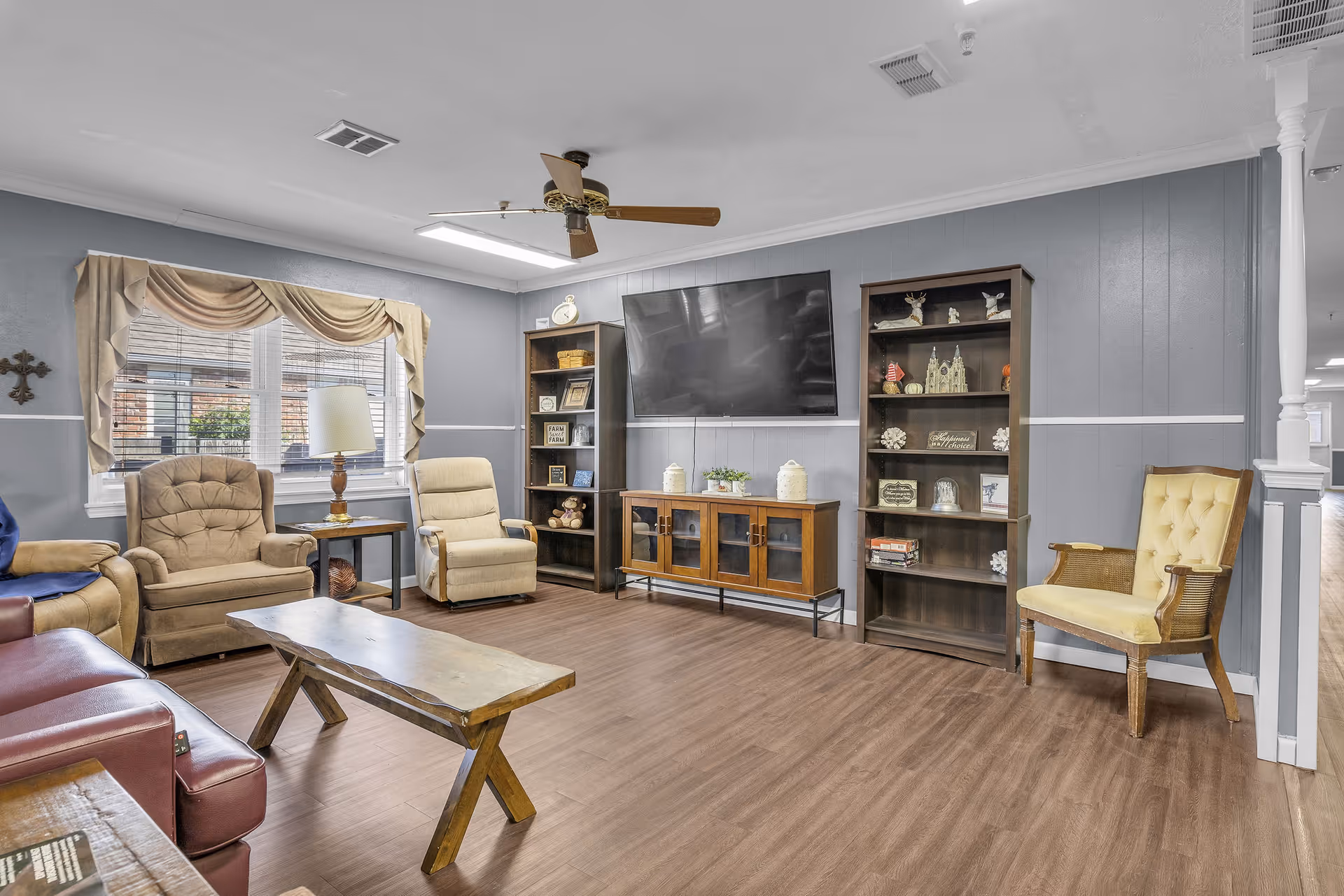 A cozy living room with a wooden floor, featuring a mounted flat-screen TV on a gray paneled wall. Below the TV is a wooden cabinet with glass doors. On either side of the TV are two tall wooden bookshelves filled with decorative items. The room has several chairs, including a beige recliner, a tufted armchair, a yellow cushioned chair, and a red leather chair partially visible. A wooden coffee table is centered in the room, and a ceiling fan with wooden blades is mounted on the white ceiling. A window with beige curtains and a table lamp is on the left side of the room.