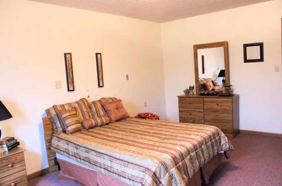 A bedroom with a double bed covered in a striped bedspread with multiple pillows. There is a wooden dresser with a mirror on the right side of the room and a nightstand with a lamp and books on the left side. The walls are plain with minimal decorations including two vertical framed pictures above the bed and a small empty frame on the wall near the dresser.