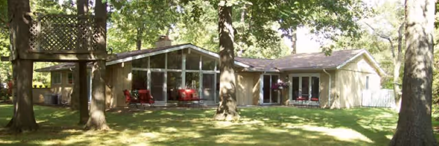 Single-story residential building with a glass-walled sunroom, surrounded by trees and red chairs on the porch.