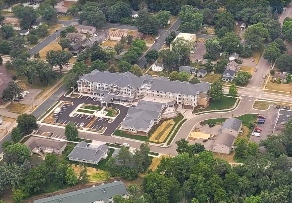 Aerial view of The Sycamore of River Falls senior living facility showing a large multi-wing building with a parking lot, surrounded by trees and residential houses.