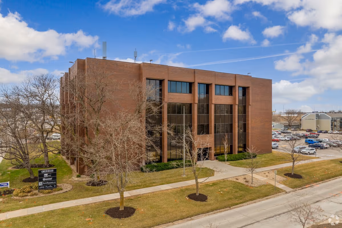 A brick, four-story building with tall glass windows, a parking lot, and bare trees.