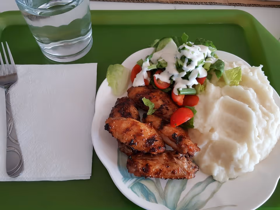 A meal on a white plate with grilled chicken, mashed potatoes, and a fresh salad topped with ranch dressing, placed on a green tray. Next to the plate is a fork on a folded white napkin and a glass of water.