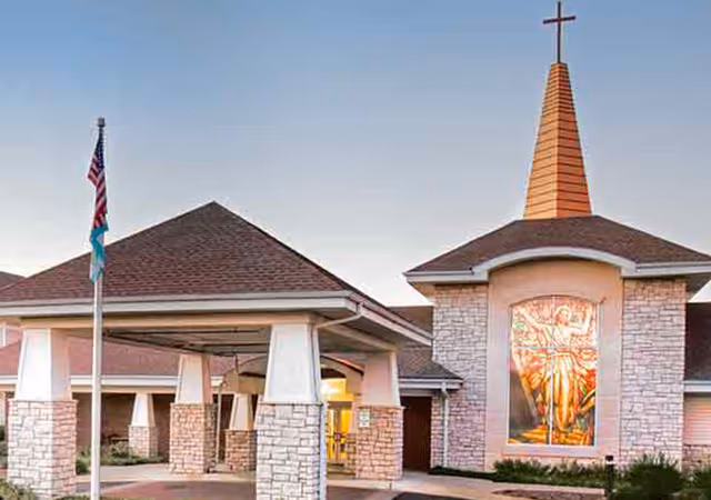 Front entrance of a senior living facility with a covered porte-cochere, stone façade, a tall steeple topped by a cross and a large stained-glass window, and flagpoles.