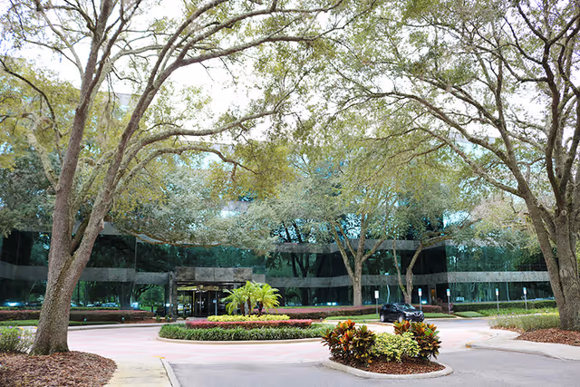 Glass-front corporate building entrance surrounded by trees and landscaped circular driveway.