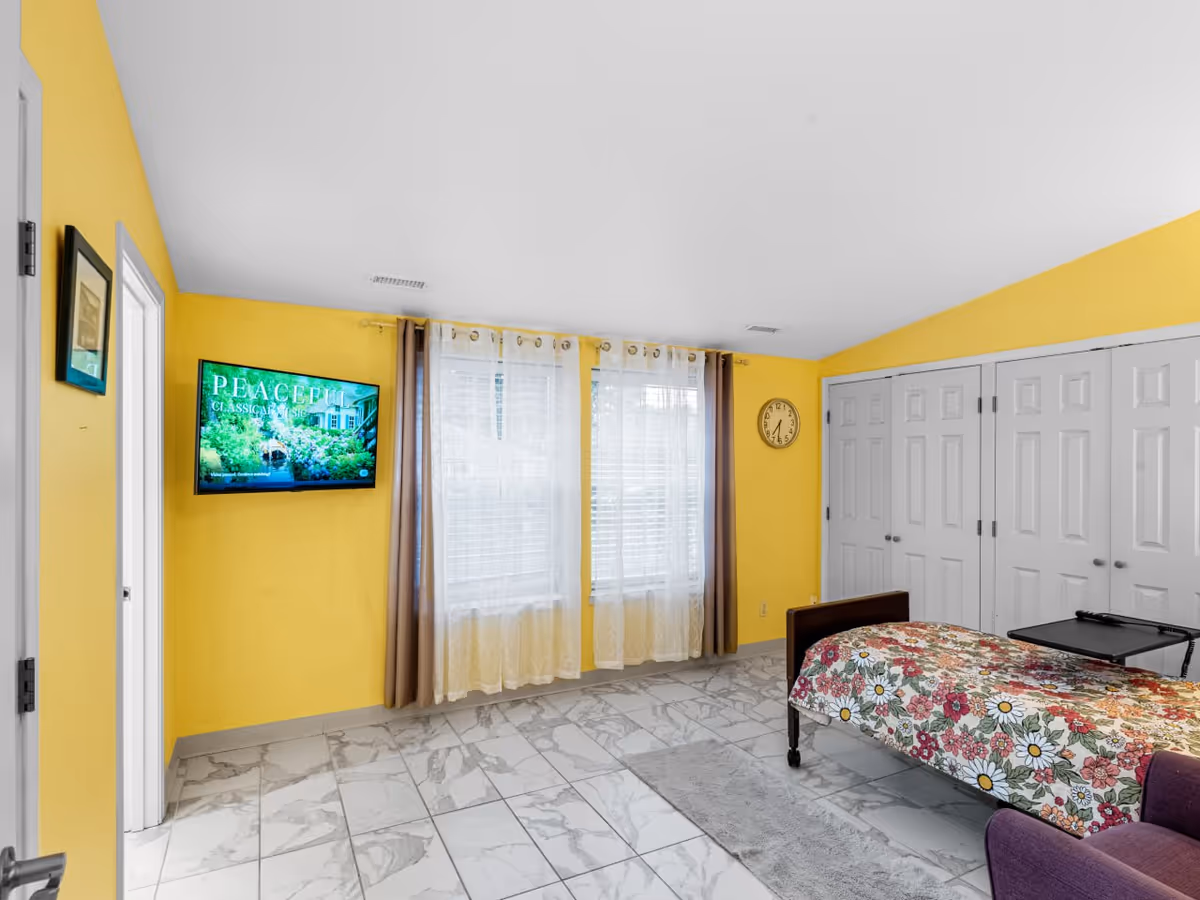 Bright yellow bedroom with floral bed, TV, windows, closets, and a purple chair.