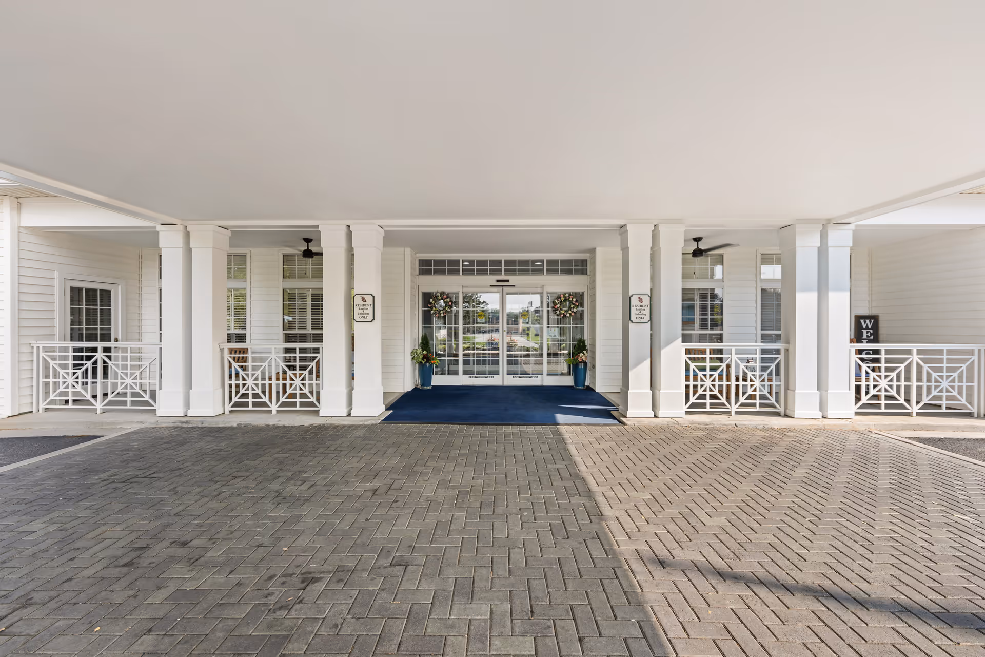 Covered porte-cochère entrance of a white senior living facility with columns, glass doors, and potted plants.