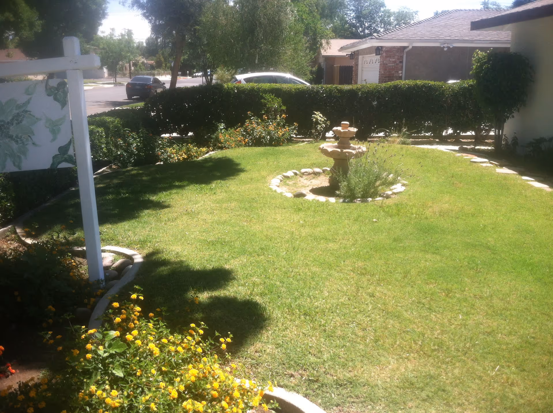 Sunlit front yard with a circular stone fountain, trimmed hedges, flower beds and a lawn in front of a single-story house.