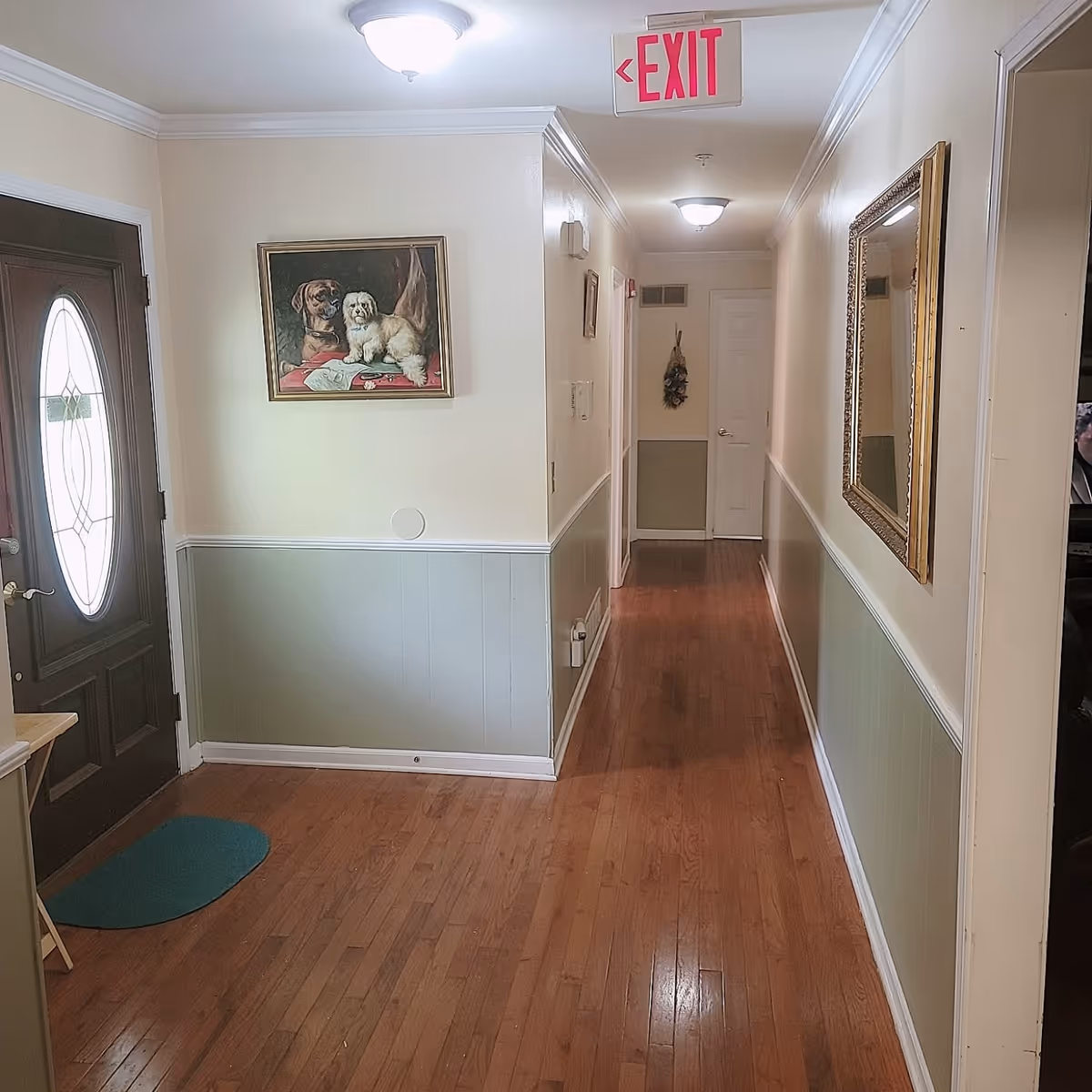 Long hallway inside Bright Rose Personal Care Home with wood floor, paintings, and exit sign.
