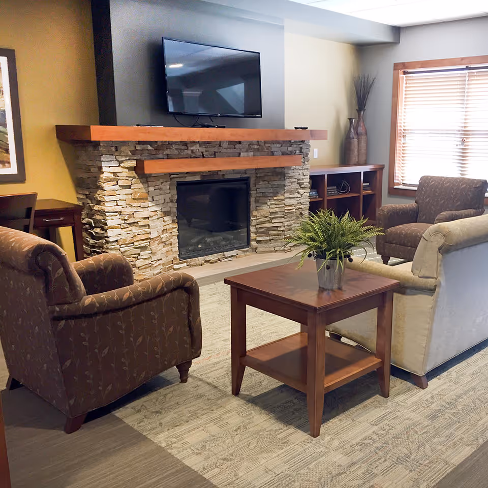 Cozy seating area with chairs and a sofa around a wooden table facing a stone fireplace with a wall-mounted TV.