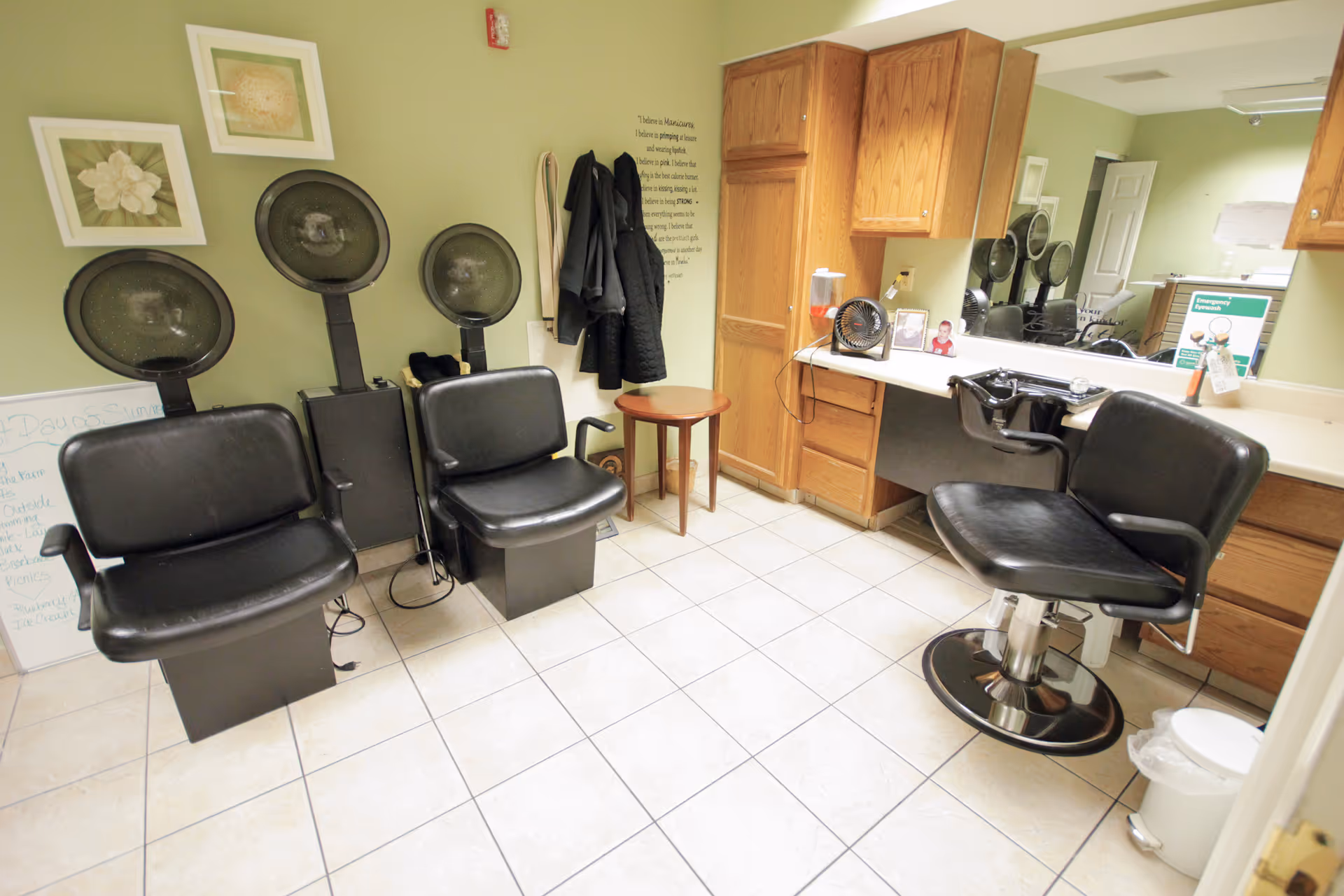 Salon area with black chairs, hair dryers, mirrors, and wooden cabinets in a green-walled room.