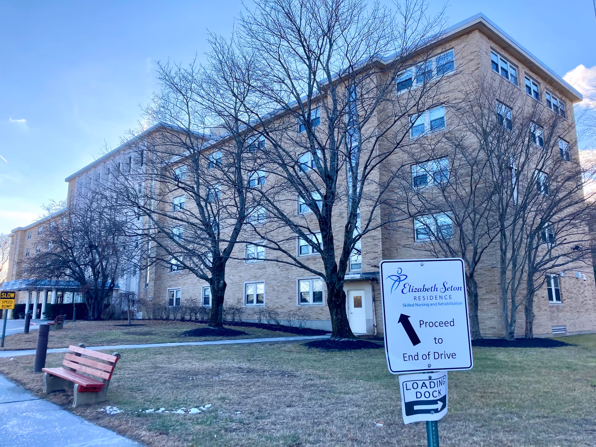 Exterior view of a multi-story brick senior residence with bare trees and a sign reading "Elizabeth Seton Residence" by the driveway.