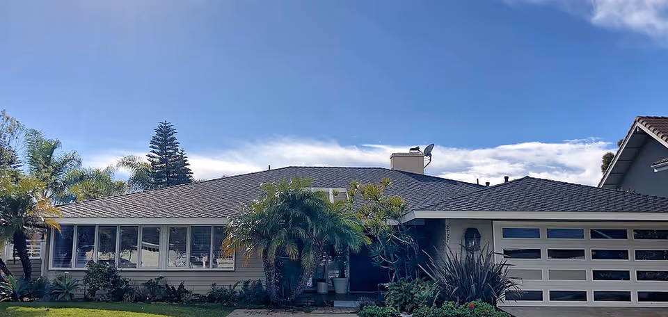 Single-story ranch-style house with a front lawn, garage, and palm trees under a blue sky.