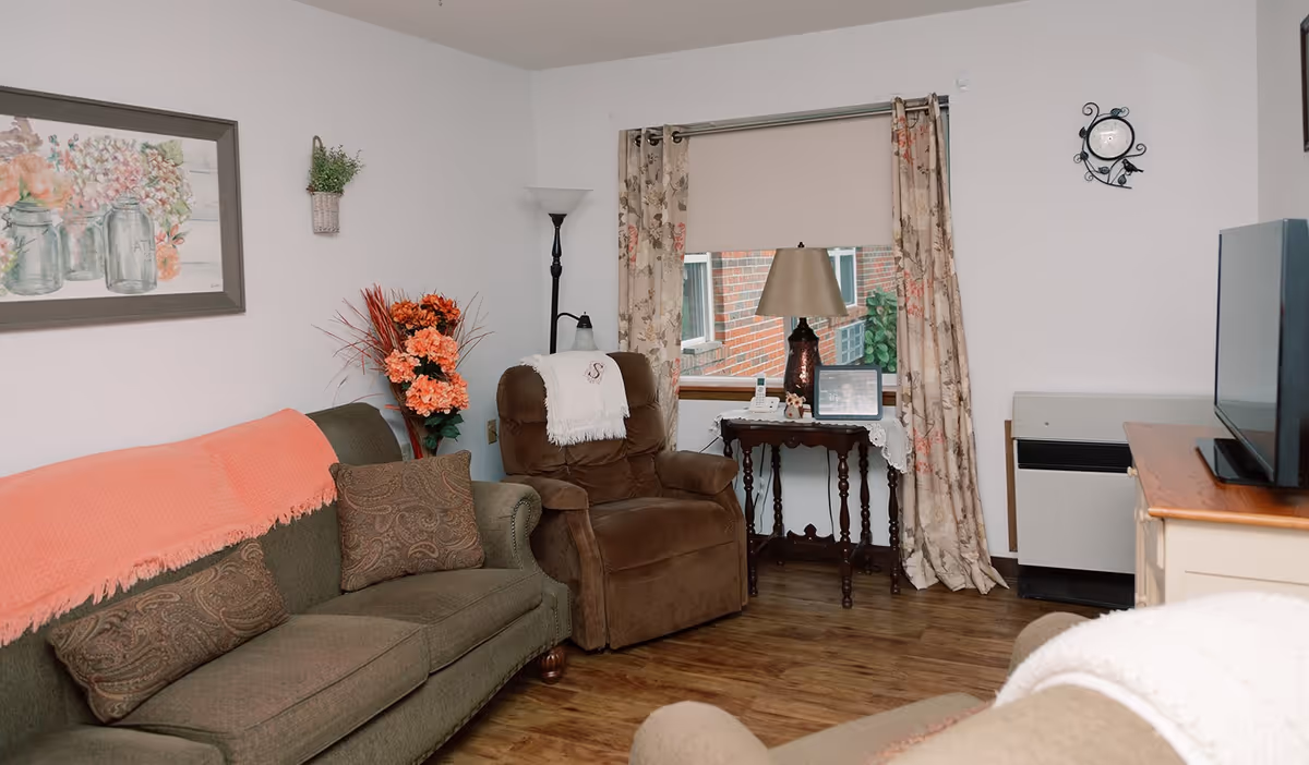 A cozy living room with a green sofa covered partially by a coral throw blanket and patterned cushions, a brown recliner with a white throw draped over the back, a wooden side table with a lamp and framed photo, floral curtains on the window, a wall clock, a floor lamp, and a TV on a wooden stand. The floor is wooden and the walls are white with a floral painting and a small wall-mounted plant.