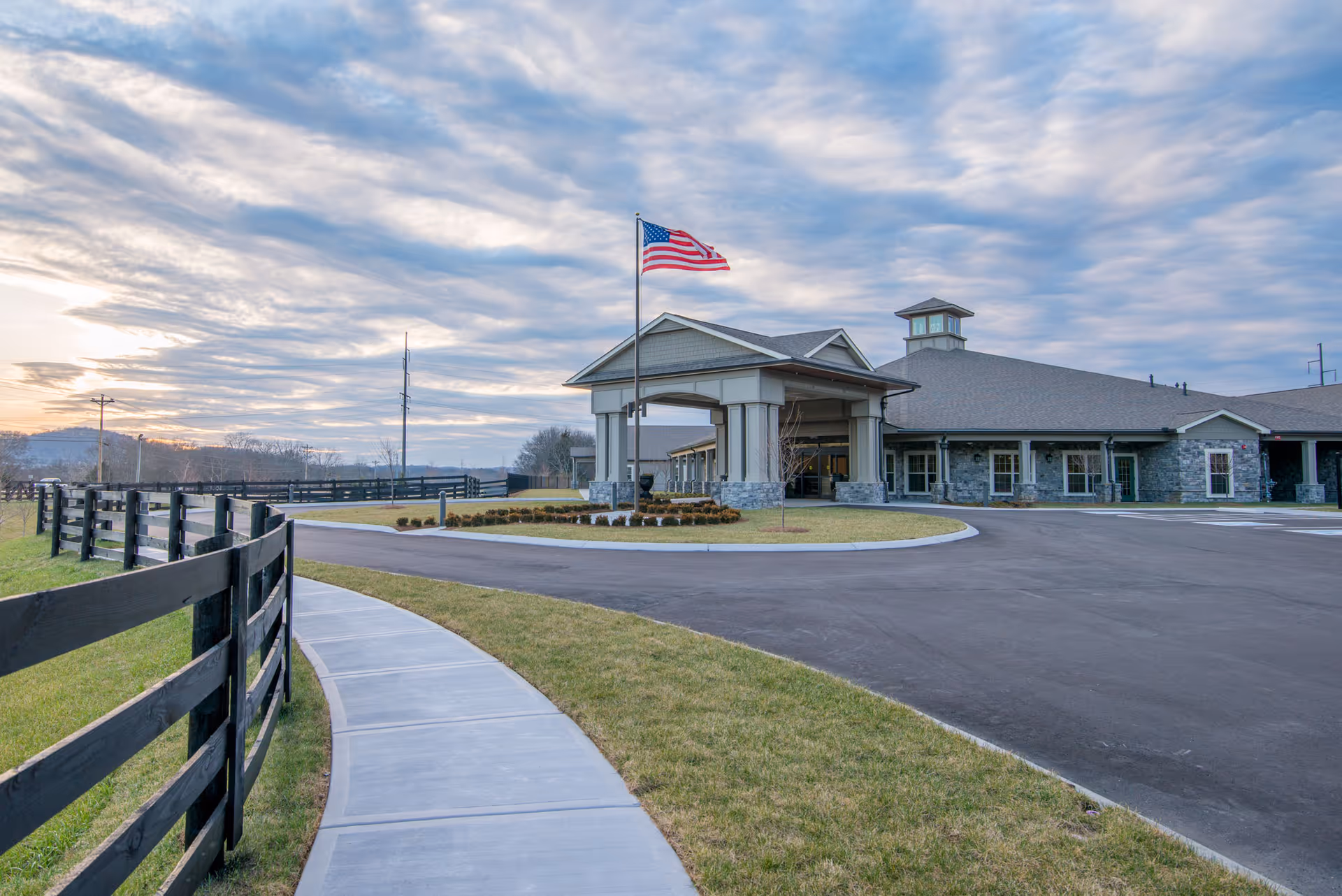 Front exterior of The Lantern at Morning Pointe facility with flag, driveway, and curved path.