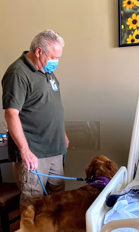 Caregiver in mask stands beside a bed with a large dog in a patient room.