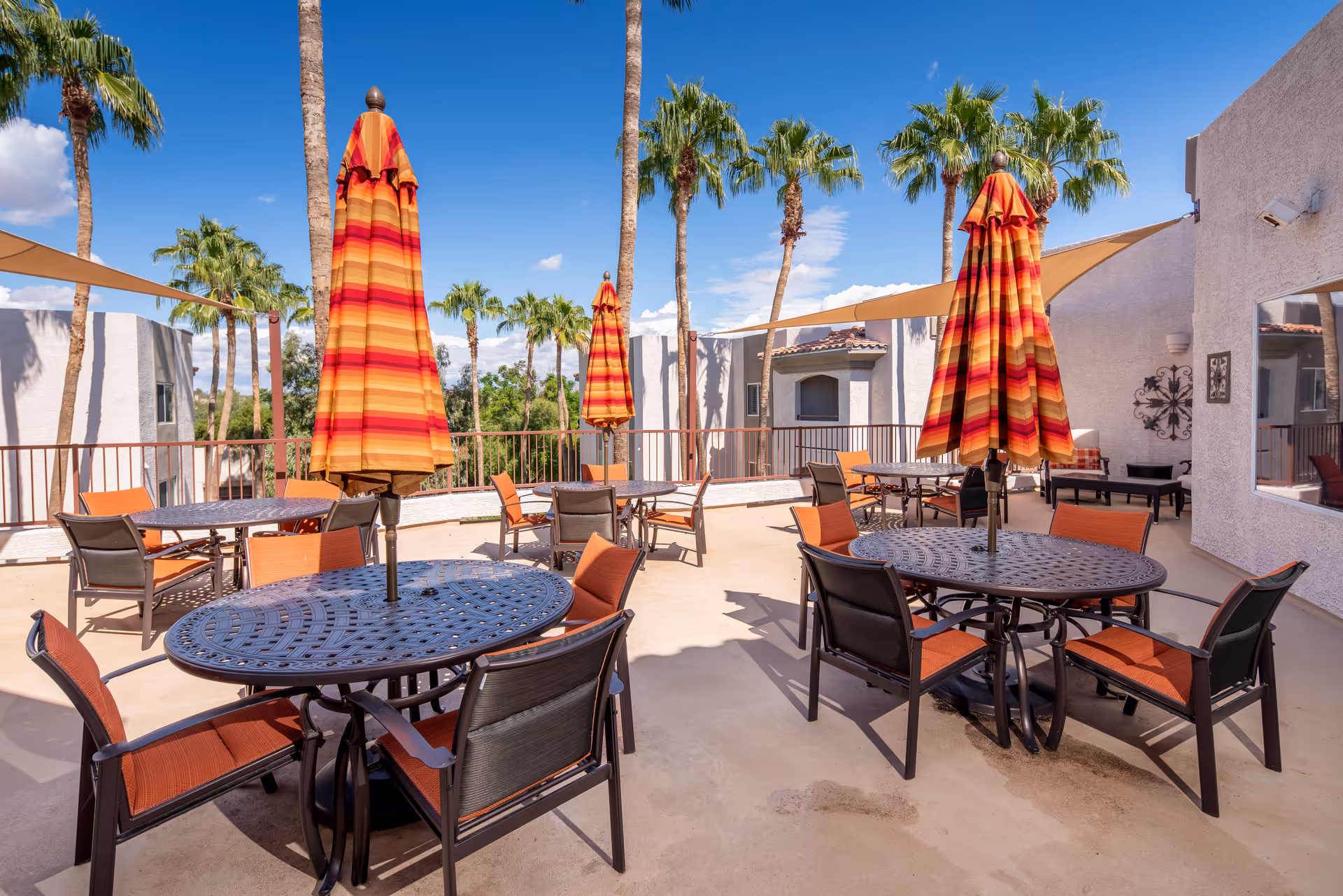 Outdoor patio with round tables, orange chairs, striped umbrellas, and palm trees.