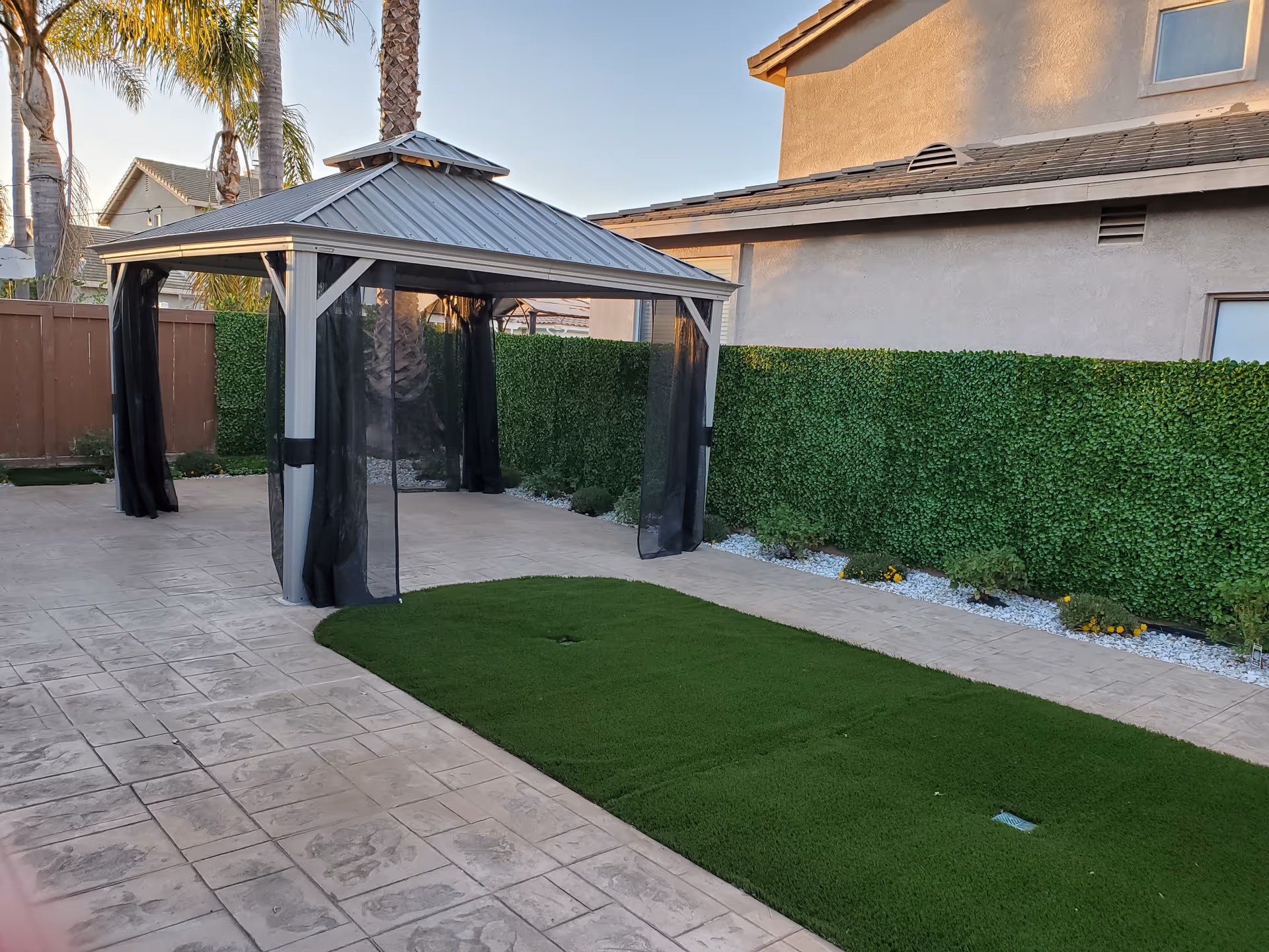 Outdoor backyard with gazebo, hedge, palm trees, and artificial turf.