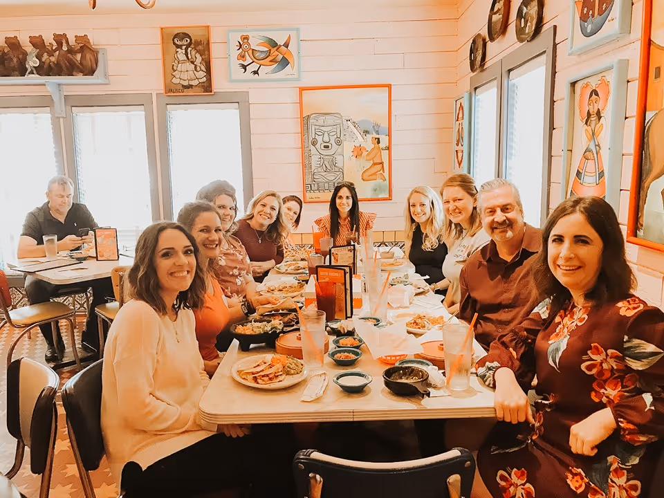 Group of adults dining together at a long table in a bright, art-filled dining room.