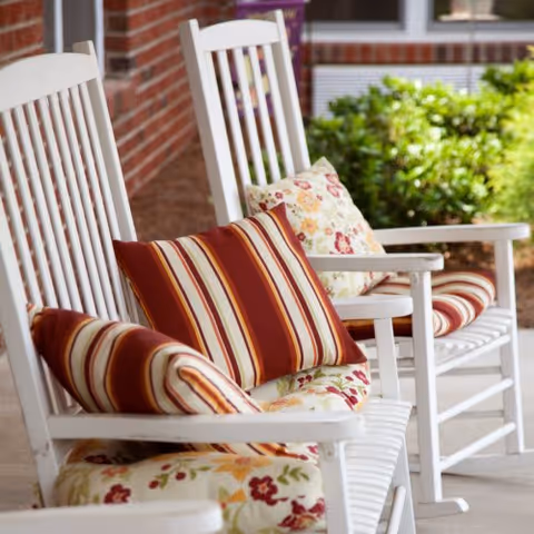 White rocking chairs with striped cushions sit on a brick porch beside green hedges.