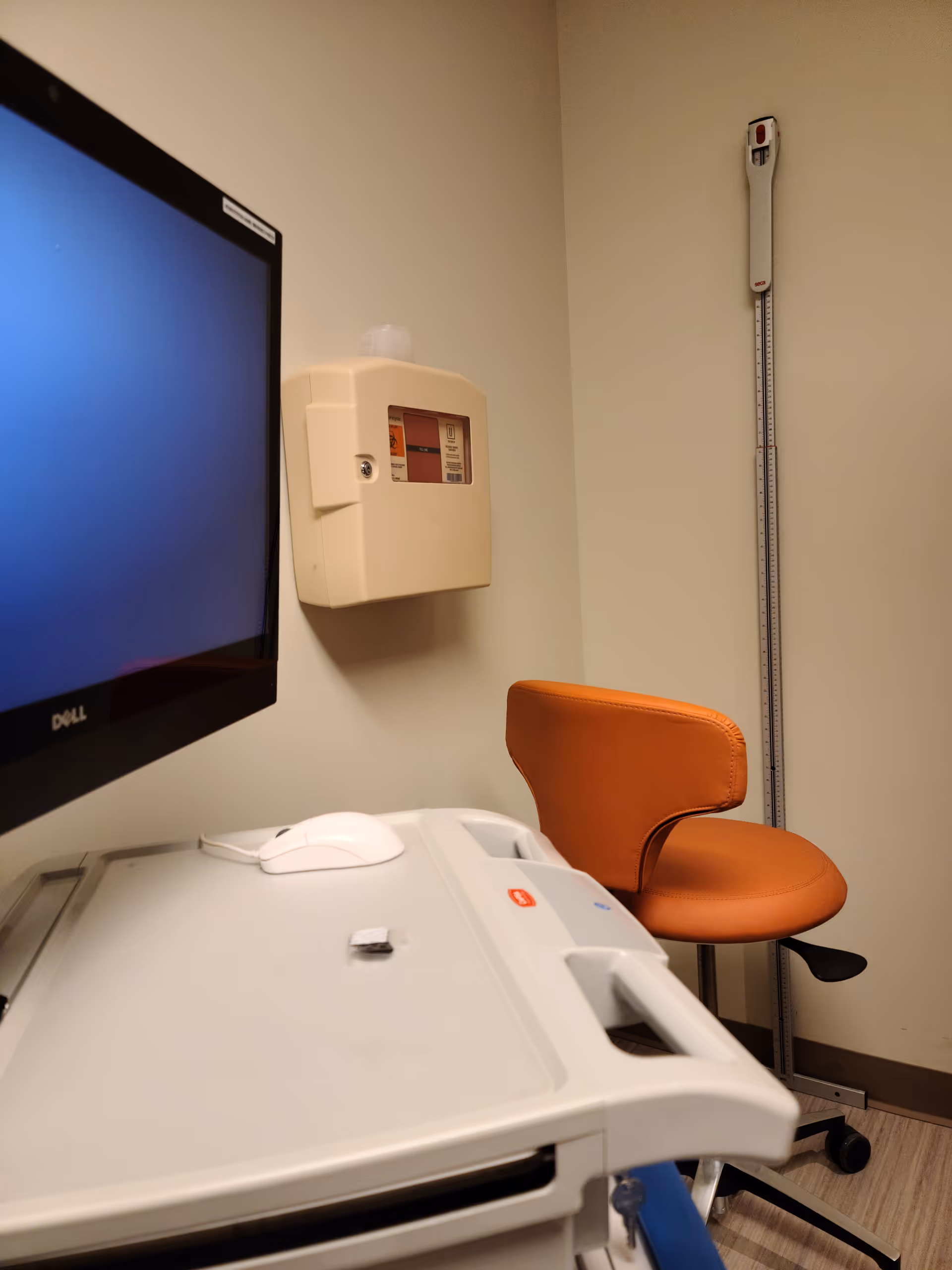 Medical examination room featuring a monitor, orange chair, and wall-mounted equipment.