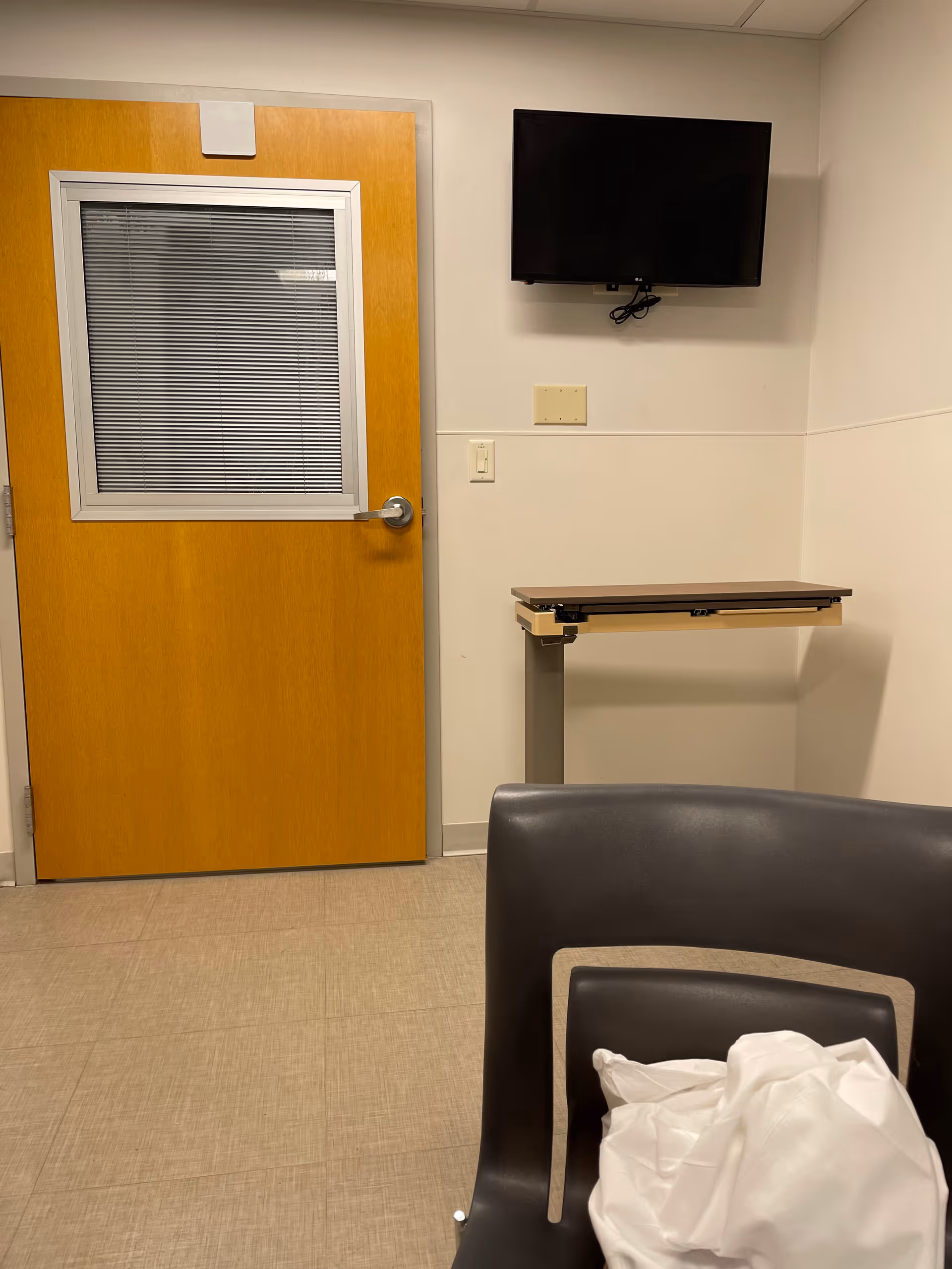 Hospital room interior with wooden door, wall-mounted TV, desk, and chair.