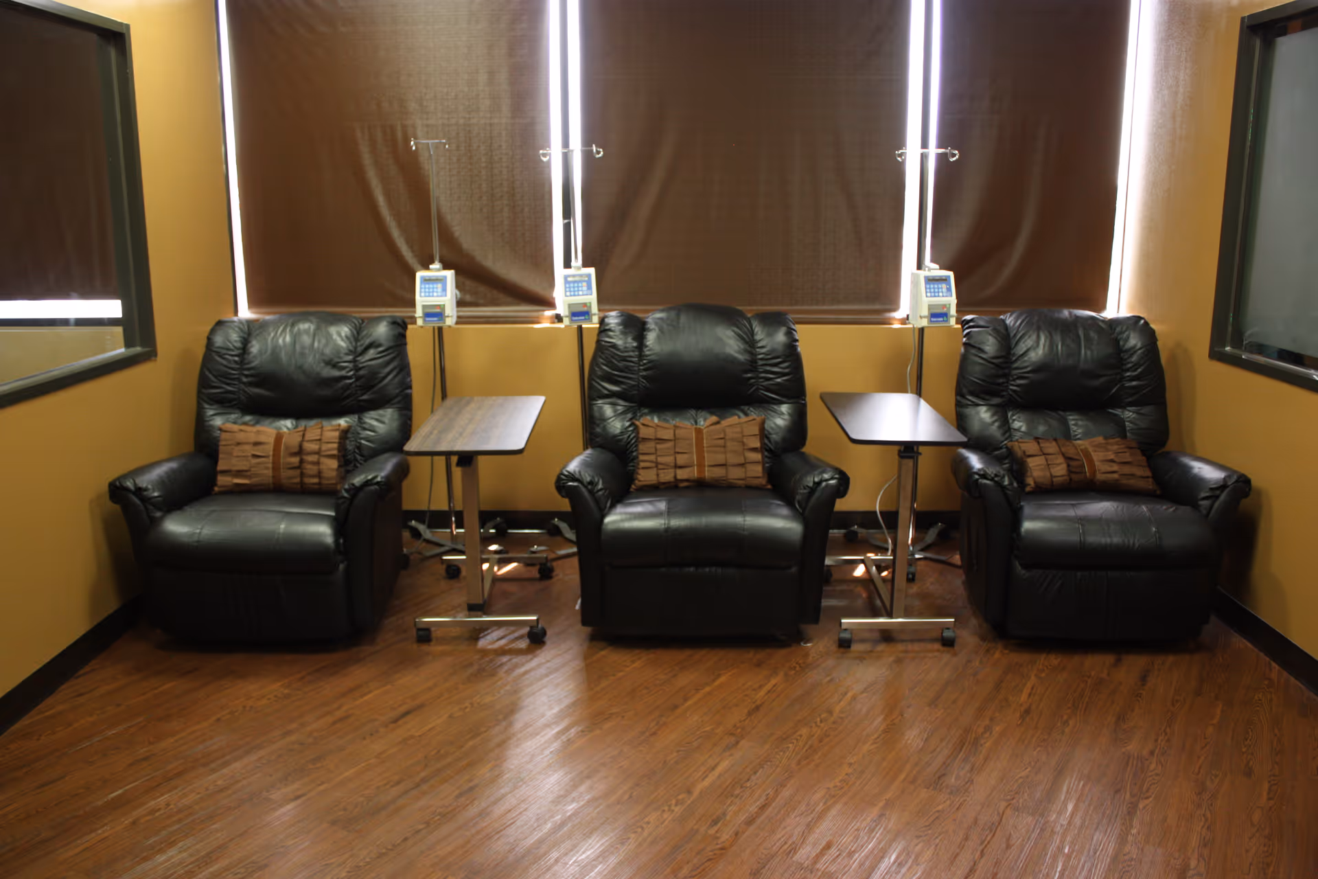 Three black leather recliners with side tables and IV stands in a tan lounge.