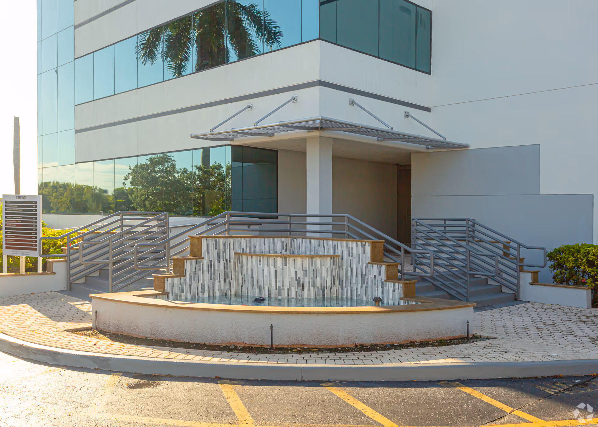 Modern glass-front building entrance with steps, ramps, and a central water feature.