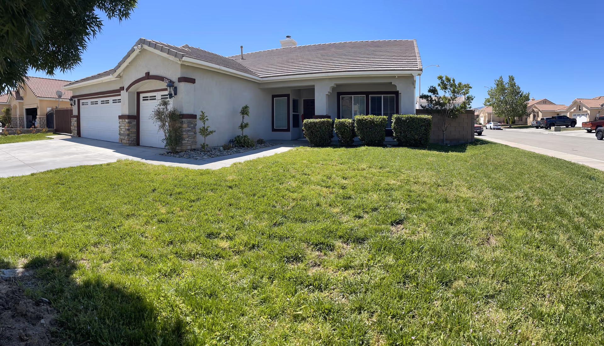 Front exterior view of a single-story house with a well-maintained green lawn, a driveway leading to a three-car garage, and trimmed bushes near the entrance under a clear blue sky.