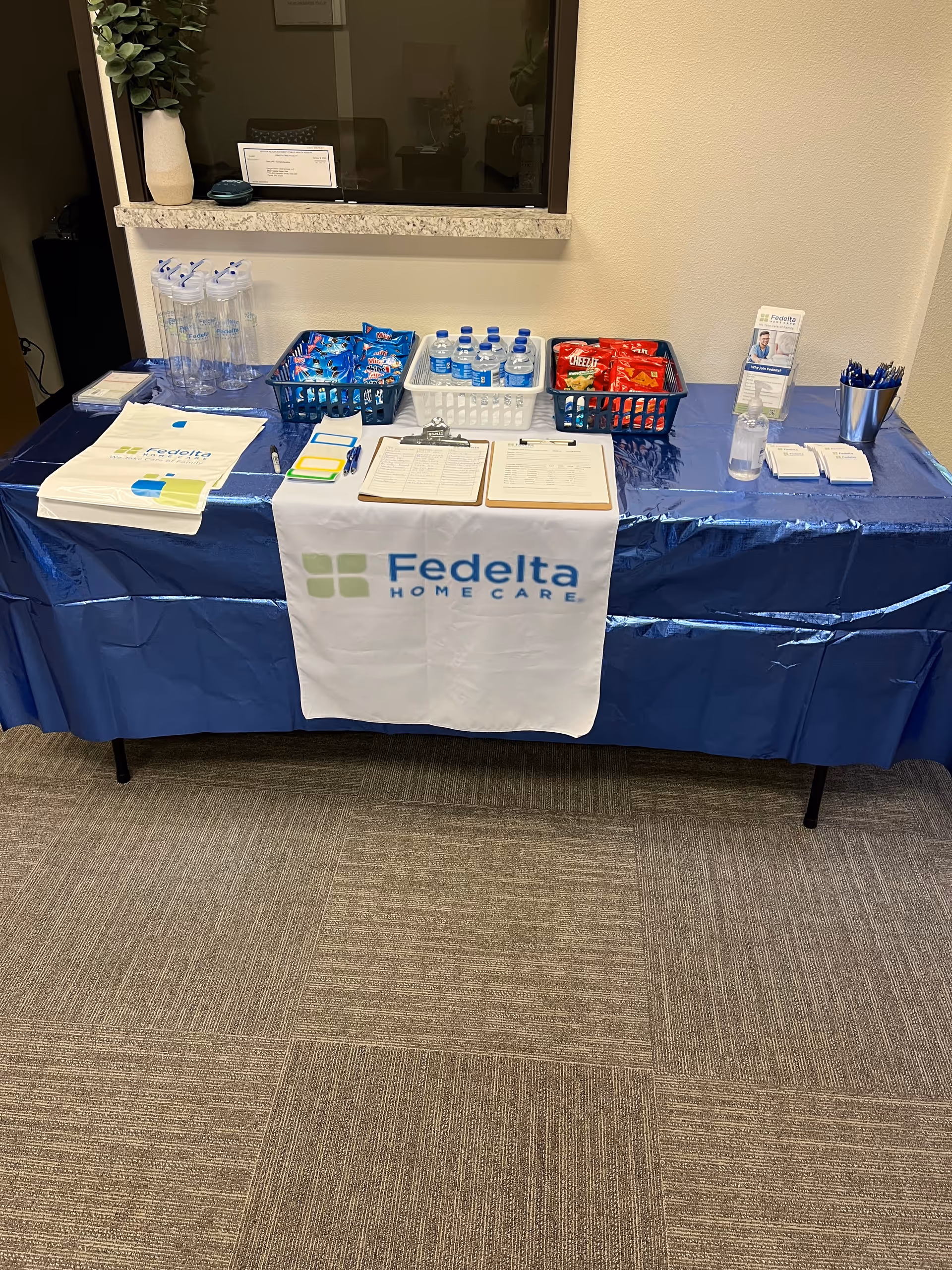 Interior table setup with blue cloth, Fedelta Home Care banner, water bottles, and snacks.