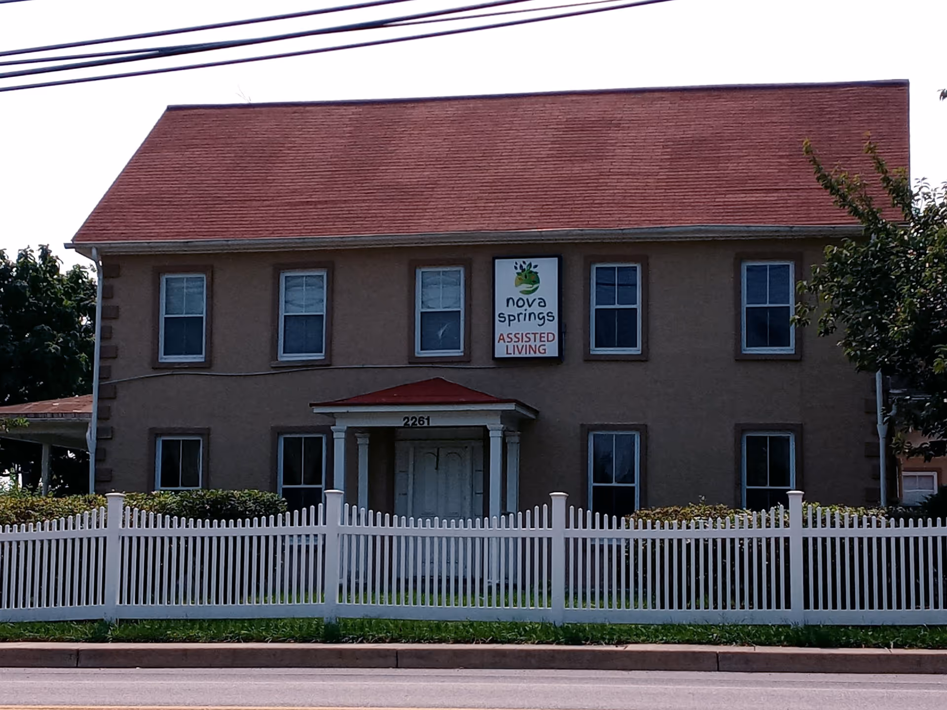 Two-story beige assisted living building with a red roof and white picket fence.