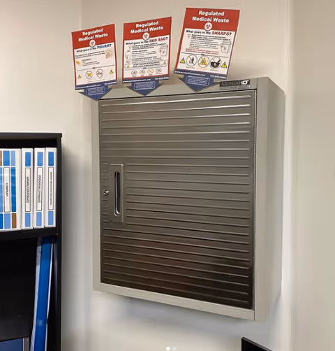 Wall-mounted metal medical waste cabinet with posters and blue file binders nearby.