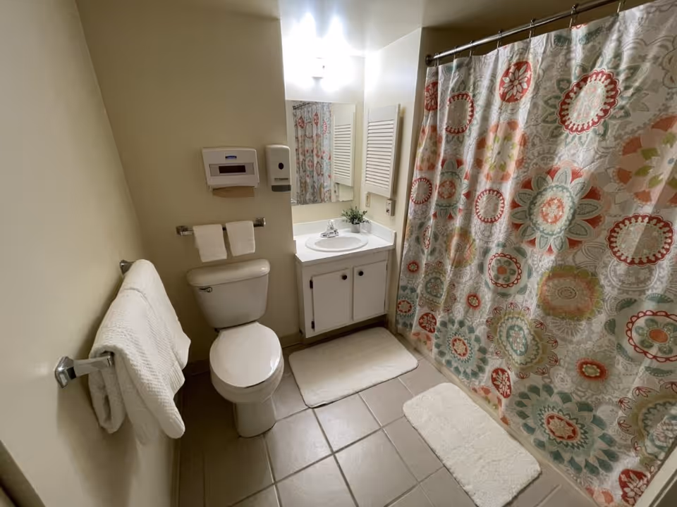 Small bathroom with toilet, sink, mirror, towel rack, and colorful shower curtain.