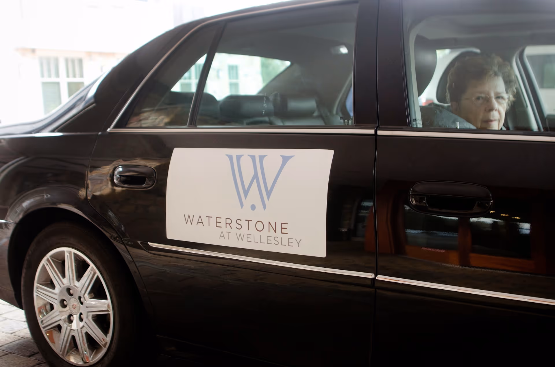 Black sedan with Waterstone at Wellesley sign on door; an elderly woman sits in back seat.